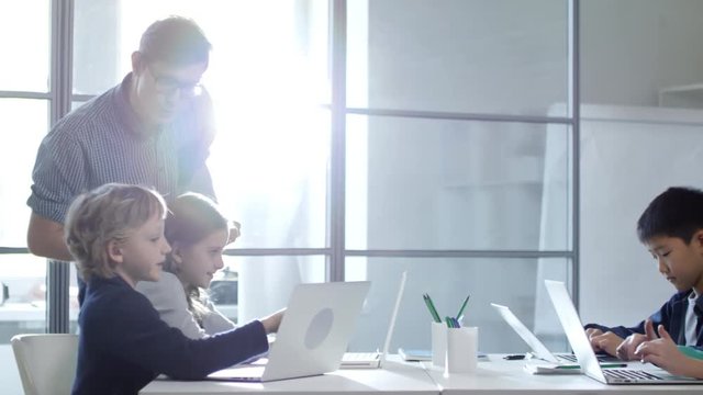 Tracking shot of four children typing something on their laptop computers in coding computer class while their teacher comes closer to each of them and checks their work