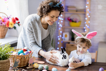 Portrait of curly blond boy wearing bunny ears  playing with white pet bunny while celebrating...