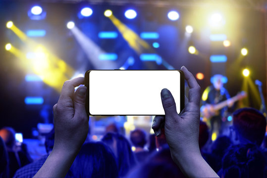 Mobile phone with isolated display in hands. Horizontal position. Live music concert in background. Crowd and lights.
