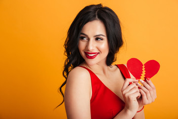 Portrait of gorgeous woman with long brown hair smiling and holding two parts of loving red paper heart, isolated over yellow background