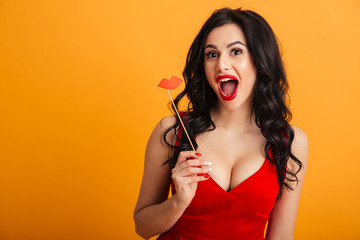 Image of joyful woman 20s in red dress smiling and holding paper lips on stick, isolated over yellow background