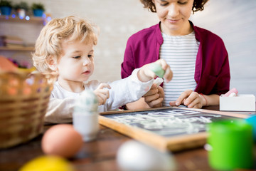 Portrait of cute little boy reaching out to write on chalkboard while making Easter decorations with mother