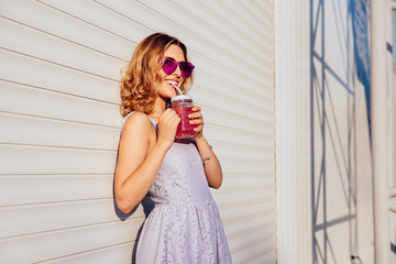 Curly cheerful girl in glamour sunglasses, drinking fresh beverage, enjoying the summer day, outdoors. Dressed in summer dress. © Maksym Azovtsev