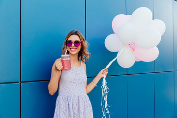 Happy beautiful woman in sunglasses drinking fresh beverage, holding a bunch of air balloons, standing against the blue wall, outdoors. Dressed in tender dress. © Maksym Azovtsev
