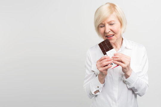 A picture of mautre lady eading a bar of milk chocolate. She likes to eat sweets. She cares about her health a lot but at the moment she wants enjoy taste of chocolate. Isolated on white background.