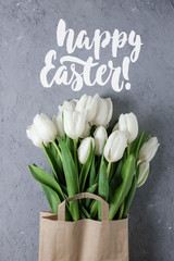 Bouquet of white tulips in a kraft paper bag with the lettering sign Happy Easter on a gray concrete background. Top view. Flat lay. Postcard for Spring Holidays.