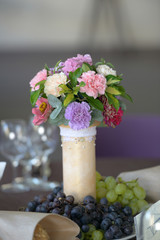 Wedding floral arrangement centerpiece with pastel colored carnations in a ivory vase decorated with pearly beads and with fresh white and red grapes positioned at its base
