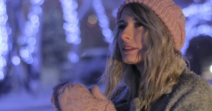 Outdoor close up portrait of a young woman in pink warm clothes trying to warm herself in night winter city. Winter concept. Christmas, winter holidays concept. Snowfall.