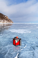 A red bag with food, a thermos with hot tea and an apple are standing on the ice of the frozen Lake Baikal on a frosty day. Winter travel, active way of life