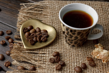 Bronze glass vase with pressed flower on a tablecloth. Golden hearth-shaped saucer with roasted coffee beans. Pure arabica on a wooden table. Lighted candle on the background. Aromatic breakfast