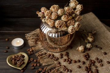 Vase with pressed flower on a tablecloth. Golden hearth-shaped saucer with roasted coffee beans. Pure arabica on a wooden table. Lighted candle on the background. Aromatic breakfast