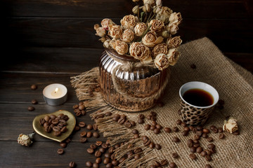 Vase with pressed flower on a tablecloth. Golden hearth-shaped saucer with roasted coffee beans. Pure arabica on a wooden table. Lighted candle on the background. Aromatic breakfast