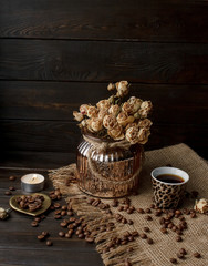 Vase with pressed flower on a tablecloth. Golden hearth-shaped saucer with roasted coffee beans. Pure arabica on a wooden table. Lighted candle on the background. Aromatic breakfast