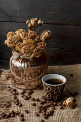 Fototapeta na wymiar Vase with pressed flower on a tablecloth. Golden hearth-shaped saucer with roasted coffee beans. Pure arabica on a wooden table. Lighted candle on the background. Aromatic breakfast