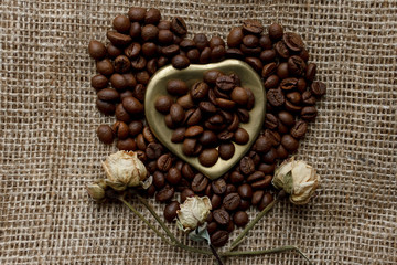 Flat lay of roasted coffee beans on a tablecloth with a golden heart shaped saucer and coffee mug. Cup of morning espresso and pressed roses. Romantic ideas
