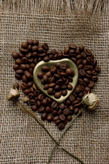 Flat lay of roasted coffee beans on a tablecloth with a golden heart shaped saucer and coffee mug. Cup of morning espresso and pressed roses. Romantic ideas