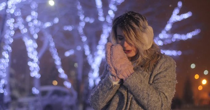 Portrait of a young woman wearing ear muffs with head tilted, trying to warm herself in night winter city. Winter concept. Christmas, winter holidays concept. Snowfall.