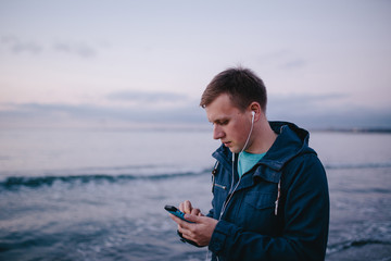 Young man in earphones holding smart phone. Young man using earphones and cellphone while walking by the sea