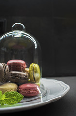 assorted color macaroon cookies on plate, glass bell cover, dark background. green pistachios, brown chocolate, pink strawberry and creme vanilla almond cakes. copyspace for text