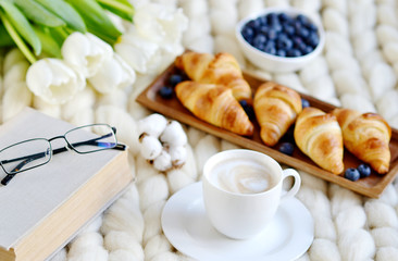 Obraz na płótnie Canvas Cup with cappuccino and croissants, berries, white pastel giant knit blanket, bedroom, flowers tulips, spring, woman day, morning concept