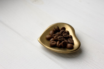 Roasted coffee beans on a golden heart shaped saucer standing on a white wooden table. Isolated aromatic arabic coffee grains. Latte and cappuccino coffee mix