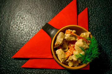 Pork in an interesting dish, baked with vegetables and decorated with dill with red napkins on a black background