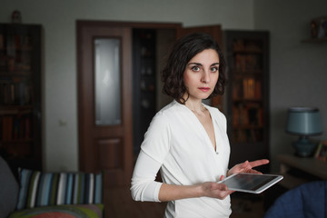 Cute young brunette woman in light clothes is in home interior. She's holding a laptop. Look at the camera.