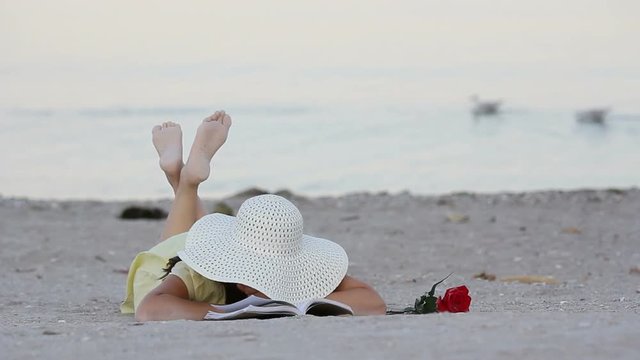 Romantic girl with a big hat laying on the beach and reading a book