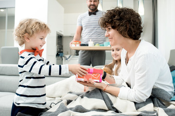 Obraz premium Side view portrait of adorable little boy giving present to his mom on Valentines day, with the rest of happy family in background