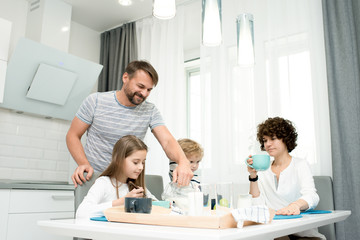 Fototapeta premium Portrait of happy young family with two children enjoying breakfast at home in modern kitchen, with father serving drinks, copy space