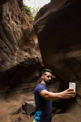 Handsome man taking selfie with cellphone in flowing shapes wall canyon in Gran Canaria, Spain. Tourist with smartphone on arid landscape. Hipster using mobile device to take photo in rocky area