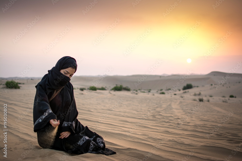 Wall mural Beautiful young woman in hijab pouring sand while sitting on desert dune during sunset.