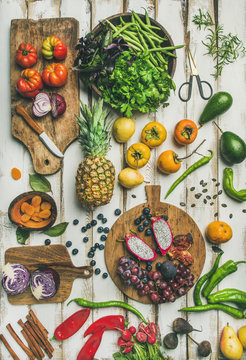 Helathy vegan food cooking background. Flat-lay of Fresh fruit, vegetables, greens and superfoods on boards over white wooden table, top view, vertical composition. Clean eating, alkaline diet concept