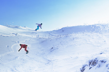 Naklejka na ściany i meble Two snowboarders ride from a high snowy mountain under a blue sky.