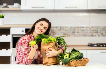 Lovely slim woman, in checked red shirt, unpacking shopping bag with green food near basket with green fruit and vegetables, concept of consuming healthy food