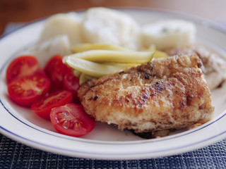 Fried white fish on a plate with tomato and green marinated cucumbers and boiled potatoes, close up.