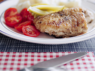 Fried portion of white fish with boiled potatoes, red mini tomatoes and pickled  cucumbers. Table knife and a napkin.