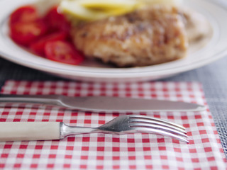 Table knife and fork on a table in focus, portion of fried white fish and red tomatoes and pickled cucumbers in the background out of focus.