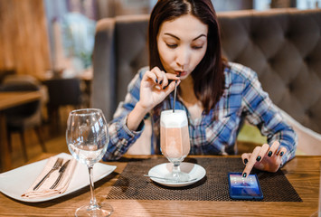 woman sitting at a wooden table in a cafe, drinking coffee and holding a smartphone.