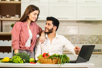 Lovely family couple spending evening together, smiling woman slicing cucumber, happy man standing nearby, using laptop and having pleasant conversation at the table with fresh green vegetables and