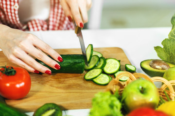 Caucasian girl with neat red manicure cutting cucumber on chopping board, wearing checked red shirt and white t-shirt, sitting at the table full of green and healthy food, closeup