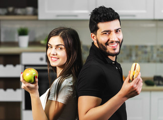 Attractive dark-haired man in black t-shirt and brunette girl hesitating between green apple and tasty hamburger, decides to eat the fresh healthy fruit, concept of choosing