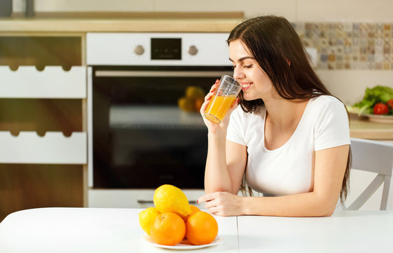 Smiling dark-haired woman having orange juice, positive relaxing atmosphere in kitchen with laptop