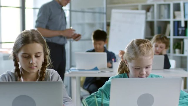 Four children of primary school age doing task on laptop computer during information technology lesson, their teacher monitoring their progress