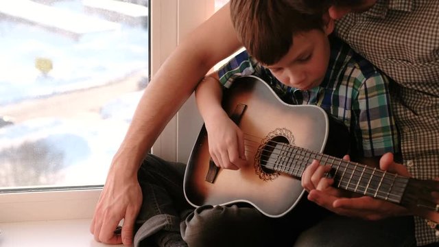 Playing a musical instrument. Dad teaches his son to play the guitar, sitting on the windowsill.