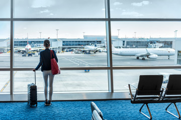 Traveler businesswoman waiting for delayed flight at airport lounge standing with luggage watching tarmac at airport window. Woman at boarding gate before departure. Travel lifestyle.