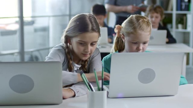 Two girls of primary school age doing task on laptop computer during information technology class, one helping another