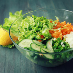Glass bowl with cutted vegetables for a salad. Close up. Bell pepper, onion, leafy greens and cucumbers cutted in pieces. Toned photo. Wooden background.