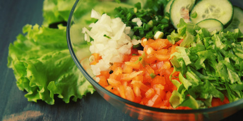 Glass bowl with cutted vegetables for a salad. Close up. Bell pepper, onion, leafy greens and cucumbers cutted in pieces. Wooden background.