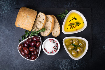 Olives, ciabatta bread, oil, herbs and spices on black stone slate background. Mediterranean snacks. Top view